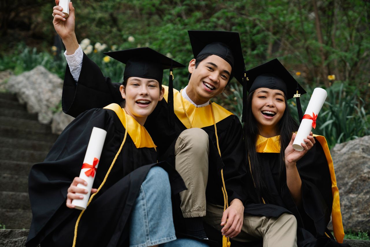 our-experience Three diverse graduates joyfully celebrating outdoors in caps, gowns, and holding diplomas.